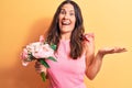 Young beautiful brunette woman holding bouquet of pink flowers over yellow background celebrating achievement with happy smile and Royalty Free Stock Photo