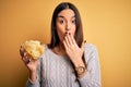 Young beautiful brunette woman holding bowl with snack potato chips over yellow background cover mouth with hand shocked with