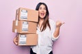 Young beautiful brunette woman holding deliver packages standing over pink background pointing thumb up to the side smiling happy Royalty Free Stock Photo