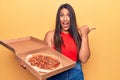 Young beautiful brunette woman holding delivery box with italian pizza pointing thumb up to the side smiling happy with open mouth Royalty Free Stock Photo