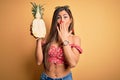 Young beautiful brunette woman holding middle pineapple fruit over isolated yellow background cover mouth with hand shocked with Royalty Free Stock Photo