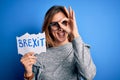 Young beautiful brunette woman holding paper with brexit message over blue background with happy face smiling doing ok sign with Royalty Free Stock Photo