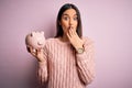 Young beautiful brunette woman holding piggy bank saving money for retirement cover mouth with hand shocked with shame for Royalty Free Stock Photo