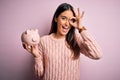 Young beautiful brunette woman holding piggy bank saving money for retirement with happy face smiling doing ok sign with hand on Royalty Free Stock Photo