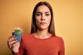 Young beautiful brunette woman holding small cactus pot over yellow background with a confident expression on smart face thinking Royalty Free Stock Photo