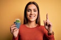 Young beautiful brunette woman holding small cactus pot over yellow background surprised with an idea or question pointing finger Royalty Free Stock Photo