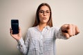 Young beautiful brunette woman holding smartphone showing screen over white background pointing with finger to the camera and to Royalty Free Stock Photo