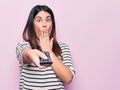 Young beautiful brunette woman holding television remote control over pink background covering mouth with hand, shocked and afraid Royalty Free Stock Photo