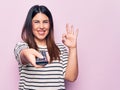 Young beautiful brunette woman holding television remote control over pink background doing ok sign with fingers, smiling friendly Royalty Free Stock Photo