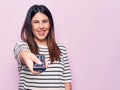 Young beautiful brunette woman holding television remote control over pink background looking positive and happy standing and Royalty Free Stock Photo