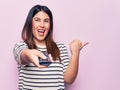 Young beautiful brunette woman holding television remote control over pink background pointing thumb up to the side smiling happy Royalty Free Stock Photo