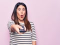 Young beautiful brunette woman holding television remote control over pink background scared and amazed with open mouth for Royalty Free Stock Photo