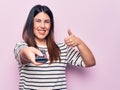 Young beautiful brunette woman holding television remote control over pink background smiling happy and positive, thumb up doing Royalty Free Stock Photo