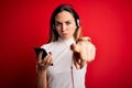 Young beautiful brunette woman listening to music using smartphone and headphones pointing with finger to the camera and to you, Royalty Free Stock Photo