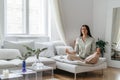 Young beautiful brunette woman meditates while sitting in yoga pose lotus sitting on white sofa in living room