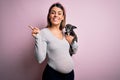 Young beautiful brunette woman pregnant expecting baby holding chihuahua puppy very happy pointing with hand and finger to the