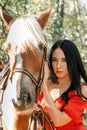 Young beautiful brunette woman in red dress posing near horse. Forest in the background Royalty Free Stock Photo