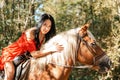 A young beautiful brunette woman in a red dress sits astride a brown horse. Summer. Sunlight