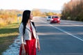 A young beautiful brunette woman in a red dress is standing on the side of the road. In the background, the road with red car goes Royalty Free Stock Photo