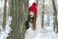 Young beautiful brunette woman in a red hat and gloves stands and snuggles up to a tree in a snowy sunny park. Portrait outdoors