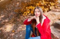 Young beautiful brunette woman with red lips and long straight hair wearing red jacket sitting outdoor in autumn park. Royalty Free Stock Photo