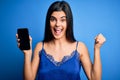 Young beautiful brunette woman wearing blue lingerie holding smartphone showing screen screaming proud and celebrating victory and Royalty Free Stock Photo