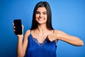 Young beautiful brunette woman wearing blue lingerie holding smartphone showing screen with surprise face pointing finger to Royalty Free Stock Photo