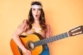 Young beautiful brunette woman wearing boho style playing classical guitar scared and amazed with open mouth for surprise, Royalty Free Stock Photo