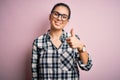 Young beautiful brunette woman wearing casual shirt and glasses over pink background doing happy thumbs up gesture with hand Royalty Free Stock Photo