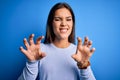 Young beautiful brunette woman wearing casual sweater standing over blue background smiling funny doing claw gesture as cat, Royalty Free Stock Photo