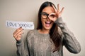 Young beautiful brunette woman wearing glasses holding paper with love yourself message with happy face smiling doing ok sign with Royalty Free Stock Photo