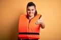 Young beautiful brunette woman wearing orange safe lifejacket over yellow background smiling friendly offering handshake as