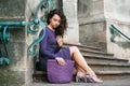 Young beautiful brunette woman wearing purple polka dot dress, holding knitted bag sitting on the stairs posing in street of city Royalty Free Stock Photo