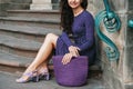 Young beautiful brunette woman wearing purple polka dot dress, holding knitted bag sitting on the stairs posing in street of city Royalty Free Stock Photo
