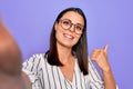 Young beautiful brunette woman wearing striped shirt and glasses making selfie by camera pointing thumb up to the side smiling Royalty Free Stock Photo