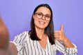 Young beautiful brunette woman wearing striped shirt and glasses making selfie by camera Smiling happy and positive, thumb up Royalty Free Stock Photo