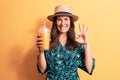 Young beautiful brunette woman wearing summer hat drinking glass of orange juice doing ok sign with fingers, smiling friendly Royalty Free Stock Photo