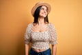 Young beautiful brunette woman wearing t-shirt and summer hat over yellow background looking away to side with smile on face, Royalty Free Stock Photo