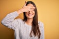 Young beautiful brunette woman wearing white casual sweater over yellow background smiling and laughing with hand on face covering Royalty Free Stock Photo