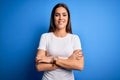 Young beautiful brunette woman wearing white casual t-shirt standing over blue background happy face smiling with crossed arms Royalty Free Stock Photo