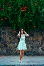 Young beautiful brunette woman in white dress stands on stone wall background with green leaves and flowers. Royalty Free Stock Photo