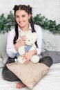 Young beautiful brunette woman in a white shirt and blue jeans sits on the bed with a pillow and teddy bear toy in his hands in Royalty Free Stock Photo