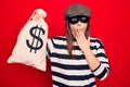 Young beautiful burglar woman wearing cap and mask holding money bag with dollar symbol covering mouth with hand, shocked and Royalty Free Stock Photo