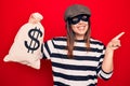 Young beautiful burglar woman wearing cap and mask holding money bag with dollar symbol smiling happy pointing with hand and Royalty Free Stock Photo