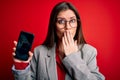 Young beautiful business woman with blue eyes holding smartphone showing screen cover mouth with hand shocked with shame for Royalty Free Stock Photo