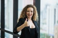 Young beautiful business woman standing near window in office. Smiling sexy lady confident in black suited showing thumbs up