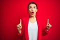 Young beautiful business woman standing over red isolated background amazed and surprised looking up and pointing with fingers and Royalty Free Stock Photo