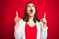 Young beautiful business woman wearing elegant jacket over red isolated background amazed and surprised looking up and pointing Royalty Free Stock Photo