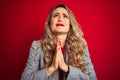 Young beautiful business woman wearing elegant jacket standing over red  background begging and praying with hands Royalty Free Stock Photo