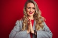 Young beautiful business woman wearing elegant jacket standing over red isolated background praying with hands together asking for Royalty Free Stock Photo